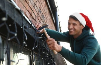A man in Santa hat standing on a ladder and decorating house with Christmas lights.