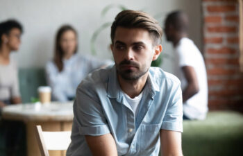 Man in denim shirt looking away, seated in a cafe. Two pairs of people converse in the blurred background.