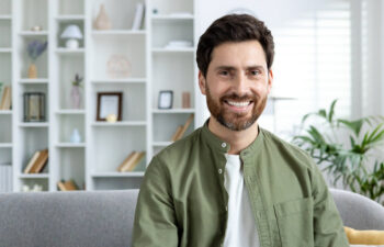 A man with brown hair and a beard, wearing a green shirt over a white t-shirt, sits on a gray couch in a modern living room with shelves in the background.
